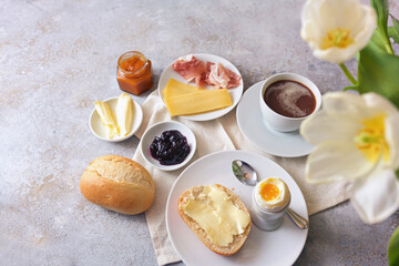 Breakfast with hearty and sweet ingredients, rolls, egg, coffee and flowers on a light stone...