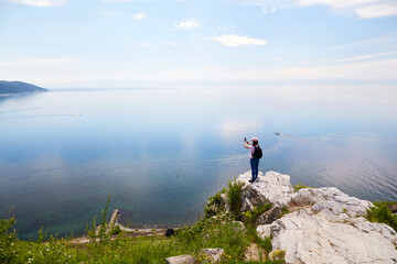 A female traveler stands on a rock and photographs Lake Baikal, the village of Listvyanka on a summer day. The concept of freedom, hiking. 