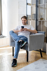 smiling man in jeans sitting in armchair with crossed legs and chatting on smartphone.