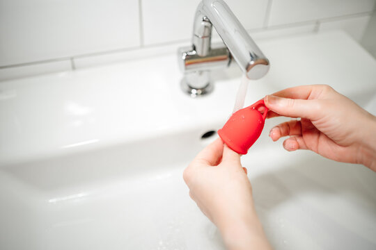 Hands of a woman washes a red menstrual cup with water under the tap at the sink. Alternative feminine hygiene product concept during menstruation. Waste-free and reusable, environmentally