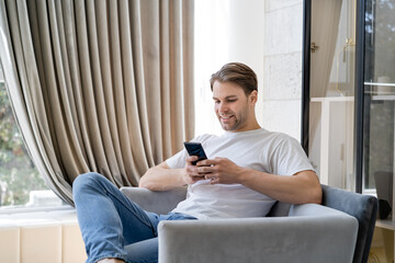 pleased man in white t-shirt messaging on cellphone while sitting in armchair.