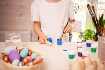 Obraz na płótnie Canvas A Caucasian boy in a white t-shirt paints eggs to celebrate the Easter holiday with bright colors. Childish art, handmade culture concept. Boy with a brush in his hand paints an egg.