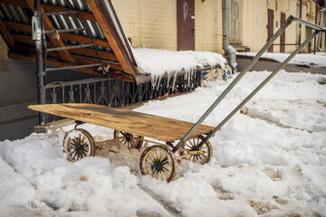 A cart-wheelbarrow in the snow near the facade of a house with icicles and a drainpipe