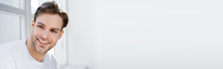 Fototapeta premium portrait of young and joyful man looking away near white wall, banner.