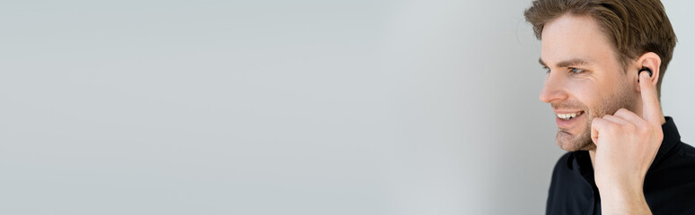 Obraz premium young man adjusting wireless earphone and smiling isolated on grey, banner.