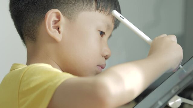 Asian boy writing a letter on a computer tablet with the electric pen in an online classroom due to Covid 19 pandemic. 5 years old kid studies at home.