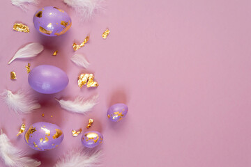 Easter eggs, violet or very peri with golden foil and white feathers on violet background, copy space.
