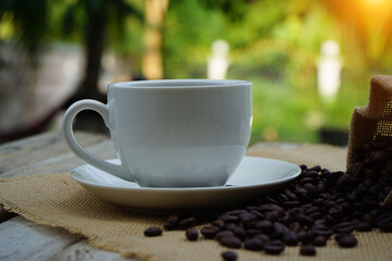 Hot Coffee cup and coffee beans roating on the wooden table beside green garden ranges in the morning sunrise