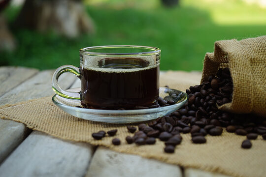 Hot Coffee cup and coffee beans roating on the wooden table beside green garden ranges in the morning sunrise