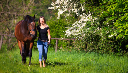 A horse and a young woman are walking across a green meadow, in the background a flowering bush..