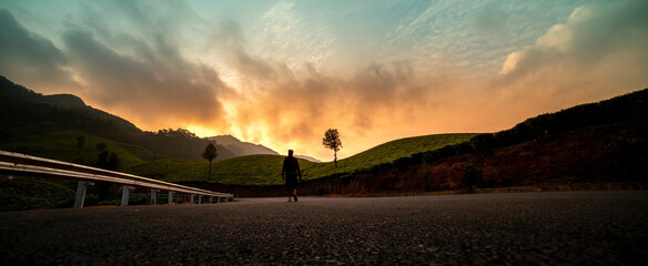 Panoramic view of wilderness scenic landscape in Munnar Kerala, Beautiful mountain sunset view and Young Photographer walking on wide road with camera  - obrazy, fototapety, plakaty