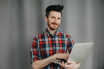 Handsome young man in a plaid shirt staying with laptop by a grey background 