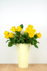 Close-up of a lot of yellow roses in a tin vase with handles on a wooden table on a neutral background. Vertical photo