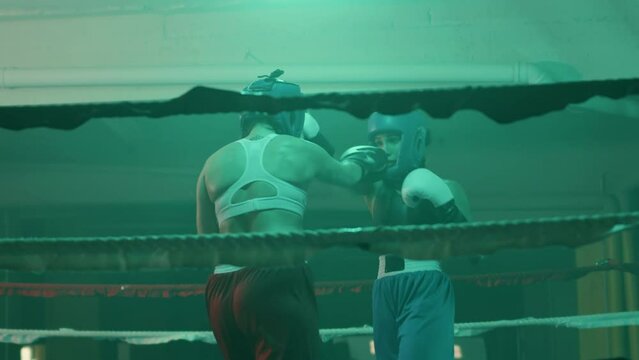 Two female boxers in helmets and boxing gloves fighting in ring at gym. Young sportswoman hitting sparring partner and knocking her out, then jumping with joy. Sport, boxing concept