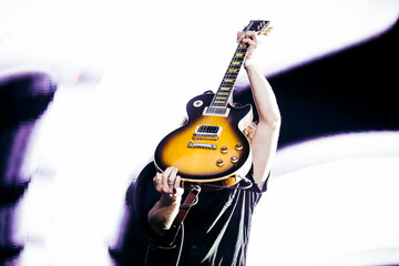 A dynamic image of a guitarist lifting a classic electric guitar above their head on stage, highlighted by a stark white background that focuses on the action and emotion of the moment.