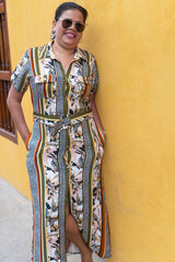 woman in a dress and sunglasses stands against a yellow wall