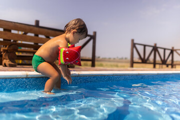 Boy with armbands playing with toys near the pool with clear water on the background of a summer sunset