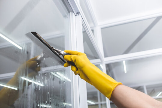 A man cleans the surface of an interior office window with a glass wiper or squeegee. The surface is sprayed with soap.