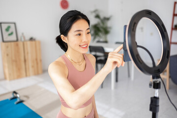 Young asian trainer woman preparing mobile phone for streaming on social media networks an online fitness video class from home - Technology, sports and healthy lifestyle.