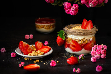 Strawberry dessert on a black background. Tiramisu and pink flowers. Strawberries with cream and cookies in a glass jar.