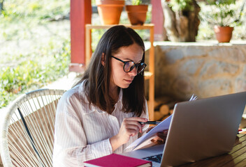 Attractive young woman wearing glasses sits at the table on the terrace and working using her laptop and hard copies of documents. - obrazy, fototapety, plakaty