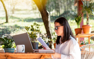 Young adult woman in glasses and white shirt works at the table with a laptop and looks through hard copies of the document. Clever female person, businesswoman. - obrazy, fototapety, plakaty