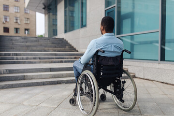 Back view of young black guy in wheelchair standing in front of stairs without ramp, having problem...