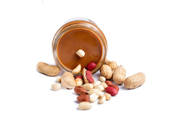 Peanut paste in a glass jar next to scattered peanuts. Isolated on a white background