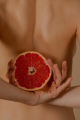 Citrus in beautiful female hands. The girl is holding a grapefruit behind her back. Juicy orange grapefruit on the background of a graceful female body. Health and fruits. Close-up