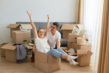 Indoor shot of happy couple having fun with cardboard boxes in new house at moving day, satisfied family with positive emotions surrounded with parcels with personal belongings.