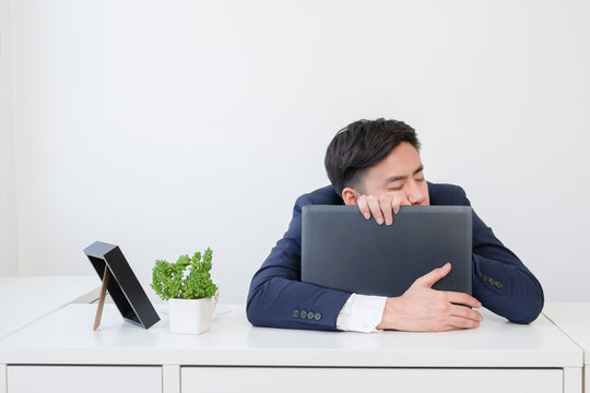 Tired young Asian businessman wear formal suit sleeping while holding laptop in arms on desk at office on break time. Business people hard working and take a nap at work