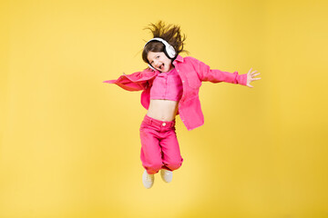 Cheerful little girl jumping and looking at camera while listening to music on headphones isolated on yellow background