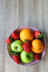 Pink bowl filled with fresh apples, oranges and strawberries on wooden table. Flat lay.