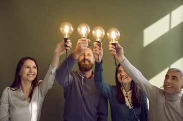 Team of smiling people raising up bright, shining Edison light bulbs as symbol of creative ideas....
