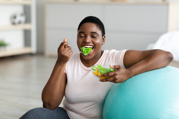 Sports and healthy eating for slimming. Plump black woman with fitness ball enjoying tasty...