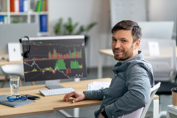 Mature man working on computer in office and smiling at camera, monitor with graphics and charts