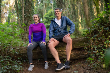 Naklejka na ściany i meble Happy sporty family resting after training in forest. Caucasian man and woman sitting on log smiling. Sport, nature, hobby concept