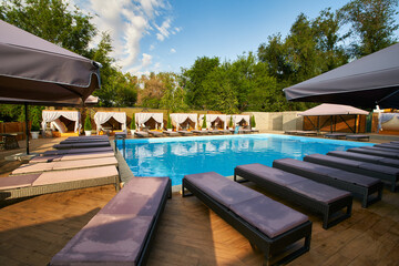 Swimming pool area with gazebos, deck chairs and sun umbrellas along the poolside in luxury resort. Summer houses surrounded with greenery and blue water. Tranquility and relax atmosphere on vacation.