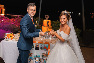 Loving couple at an international wedding at sunset. Beautiful sophisticated bride of Asian appearance. Strong manly groom of Caucasian appearance. Mixed marriages. Orange wedding cake.