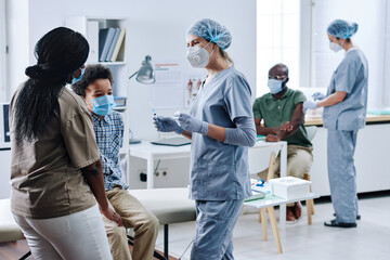 Nurse in protective wear talking to mother of little child while they visiting hospital for...