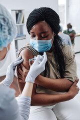 African girl in protective mask getting vaccine in her arm while sitting at hospital during pandemic
