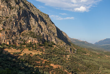 High mountains, green valley, olive trees and the mountain road. On the background is blue sky and white clouds. Greece