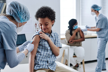 African brave boy getting vaccination in shoulder from nurse during procedure at hospital with other people in background - obrazy, fototapety, plakaty