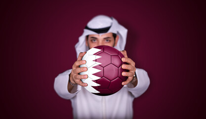 Arab man holding soccer ball in hand with Qatar flag standing on red background.