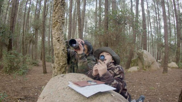 Young mother and son dressed in camouflage clothes photo hunting in forest, staying behind stone. Long-haired woman making wildlife photos while boy observing through spy pipe. Nature, leisure concept