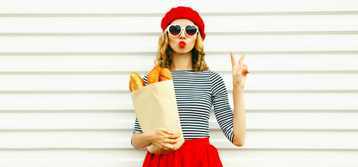Portrait of beautiful woman blowing her red lips sending sweet air kiss holding grocery shopping paper bag with long white bread baguette wearing french red beret on white background - obrazy, fototapety, plakaty