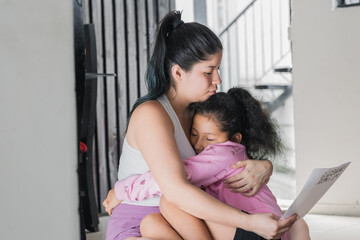 beautiful latina woman hugging her daughter while reading the letter her daughter gave her as a gift. tender moment between mother and daughter sitting on the floor of their home. concept of home. - obrazy, fototapety, plakaty