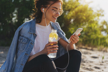 Young positive woman in optical eyeglasses smiling during a video call  on smartphone device, attractive student girl listening to music and reading new on cellphone outdoors - obrazy, fototapety, plakaty