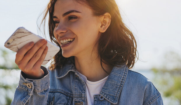 Portrait of young positive woman smiling while recording audio messages using a smartphone outdoor
