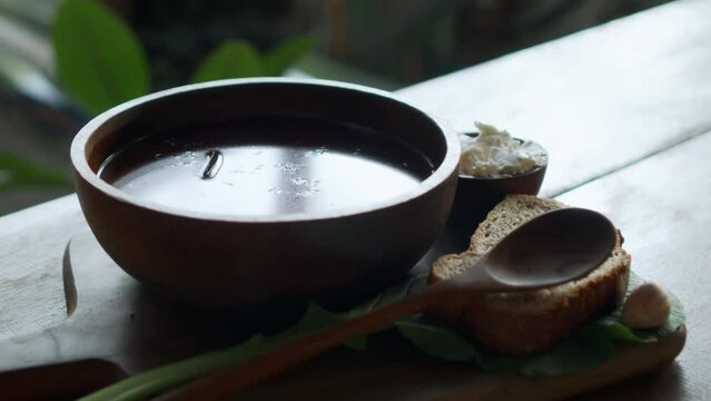 Traditional Russian fresh hot soup borsch with beetroot, red color, with sour cream and fresh bread in a wooden plate standing on a wooden table
